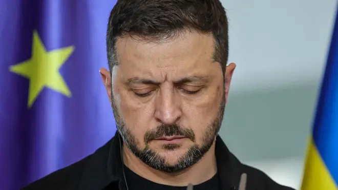 Владимир Зеленский, скорбно опустив глаза, стоит на фоне флага Евросоюза