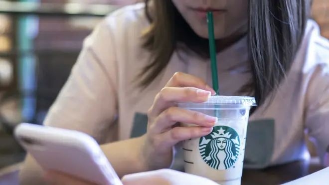 A imagem mostra, do nariz para baixo, uma pessoa branca,soccer bet procabelos lisos e castanhos na altura do ombro, segurando um copo transparente com o símbolo da Starbucks e um canudo verde levado à bocasoccer bet prouma mão. Na outra mão, um celular na horizontal.