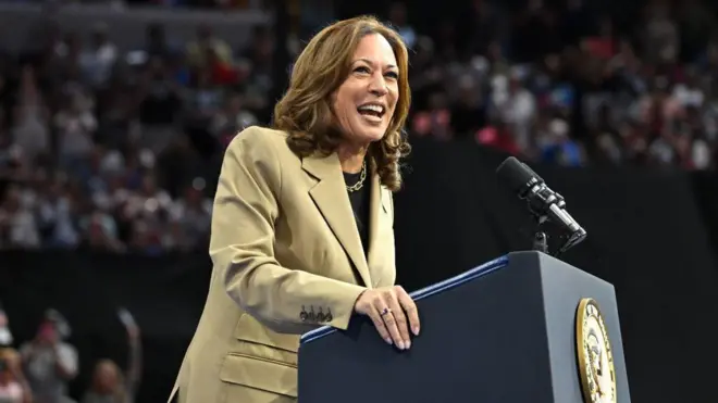 A imagem mostra Kamala, uma mulher morena,bets sports apostascabelos na altura dos ombros, vestida com um blazer bege, falandobets sports apostasum púlpito. 