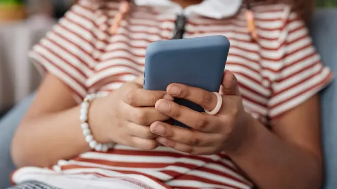 Una niña con una camiseta de rayas rojas y blancas lleva una pulsera de cuentas y un anillo rosa y sostiene un teléfono móvil con funda azul.