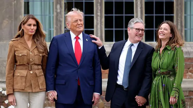 Donald Trump ve Keir Starmer eşleriyle el ele tutuşmuş, Starmer'ın eli Trump'ın omzunda ve gülümsüyorlar