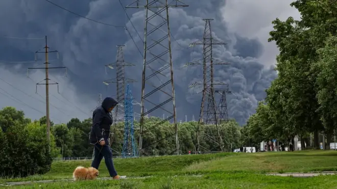 Una mujer pasea su perro por un parque y de fondo se ven las columnas de humo negro de la refinería atacada por Ucrania.