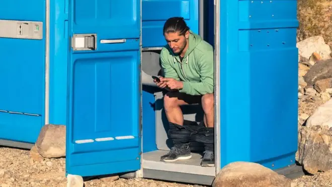 A man sitting on the toilet in a portable toilet in the middle of the Death Valley desert, in the United States. He has his trousers rolled around his ankles and is reading his phone.  The toilet door is wedged open by a rock and another portable toilet is next to hium, with the door closed. Rocks are scattered around and there are barren hills in the background.