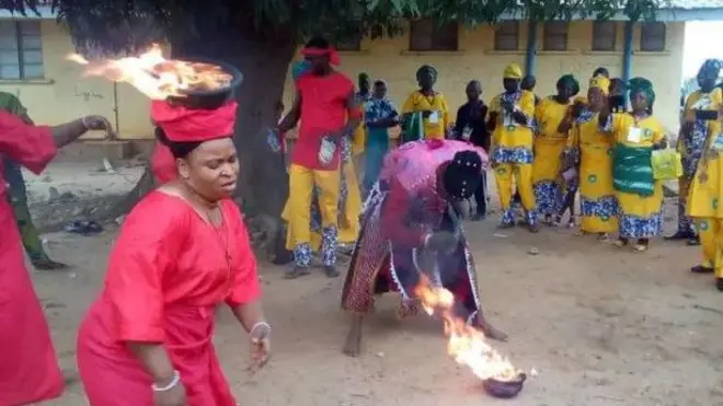 Awọn Onisango wọ asọ pupa, ru ina sori, ọkunrin kan to di irun sori n da ina sinu ikoko nigba ti awọn to wọ ankara alawọ Yẹlo n woran