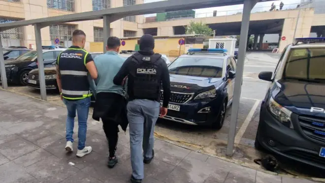 Un hombre con camiseta azul de manga larga flanqueado por dos agentes de Policía protegidos por chalecos balísticos. Caminan hacia autos policiales estacionados. 