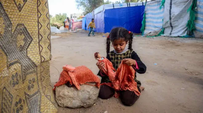 طفلة تحمل دمية خارج خيمة، حيث لجأت عائلات نازحة بسبب الحرب، في دير البلح في وسط قطاع غزة في 12 يناير/كانون الثاني 2025. الصورة التقطت نهاراً وخلف الطفلة مجموعة من الخيم.