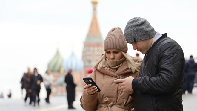Мужчина и женщина, одетые в теплые куртки и вязаные шапки, стоят на Красной площади на фоне собора Василия Блаженного и смотрят в свои телефоны