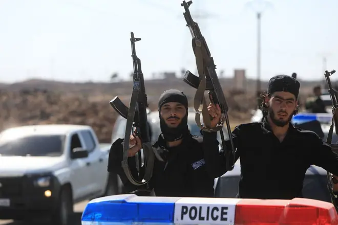 أفراد أمن سوريون يحملون بنادق وأسلحة نارية، بينما يقفون داخل سيارة كُتب على أعلاها كلمة "شرطة" باللغة الإنجليزية.