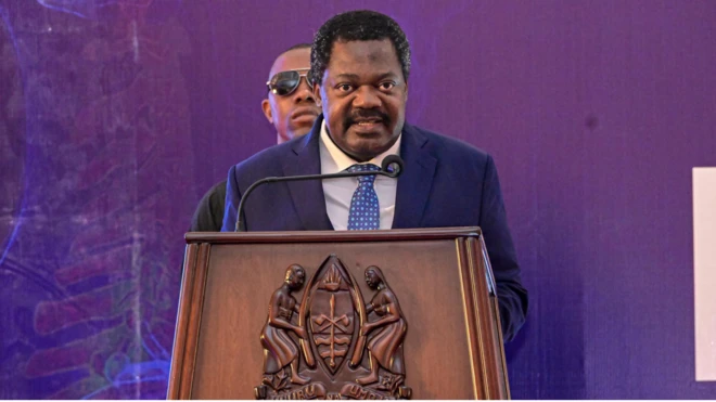 Emmanuel John Nchimbi arimo gushikiriza ijambo. Yambaye ikote ry'ubururu. Inyuma yiwe hahagaze umuntu yambaye amarori amucungereye umutekano. (Ifoto mbarankuru yo mu bubiko)