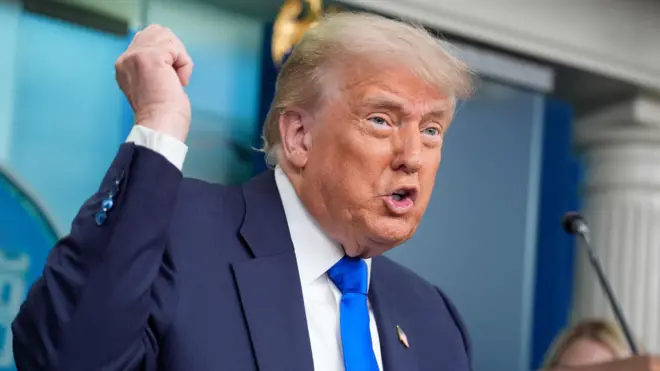 Donald Trump wears a blue suit and tie with a hand raised in the White House briefing room