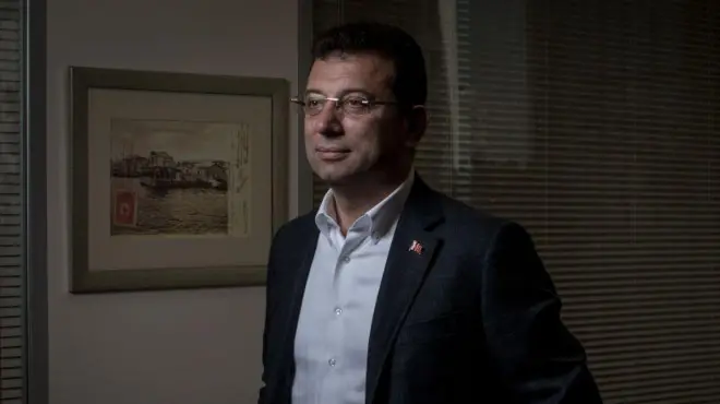 Ekrem İmamoğlu üzerinde bir takım elbiseyle, kravatı olmadan loş bir ofiste kadrajın solundan uzaklara bakıyor