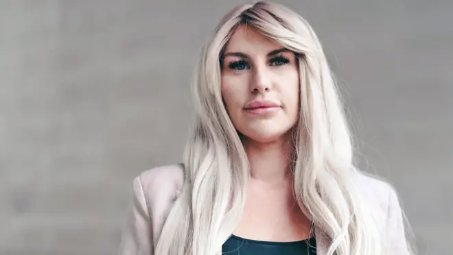 Rebecca posa diante de um muro de pedra clara. Ela tem cabelos loiros longos e ondulados, veste uma blusa preta e uma jaqueta rosa-clara e olha para a câmera com expressão pensativa