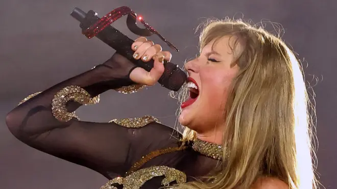 Taylor Swift on stage side on, singing into a microphone with her mouth fully open and eyes closed