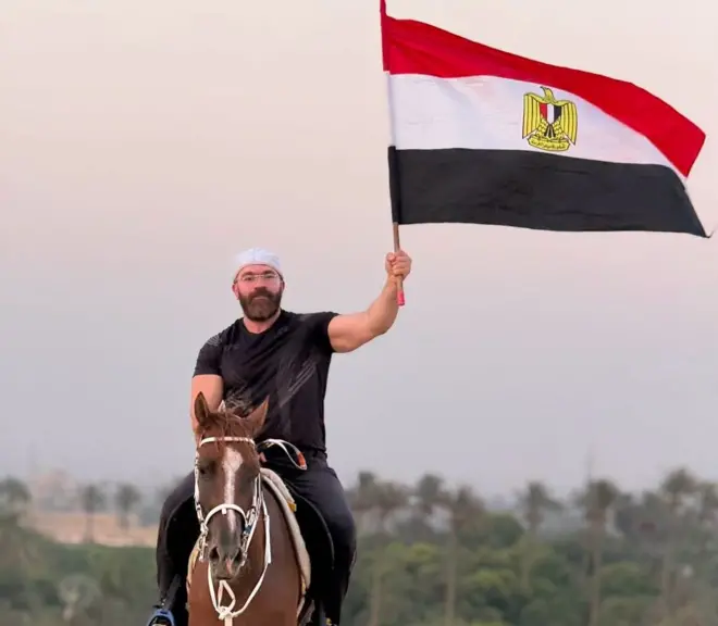 المدون المصري، رامي عادل يمتطي حصاناً ويرفع بيده اليسرى علم مصر، وحوله رمال وأشجار.