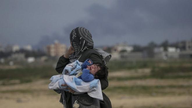 黒煙の立ち上るガザ地区を、赤ちゃんを抱えて移動する女性