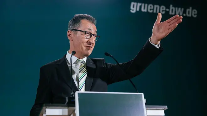 Mikrofon başında gözlüklü bir adam gülümseyerek eliyle kalabalığa işaret ediyor. Arkada Yeşiller Partisi'nin logosu var. 