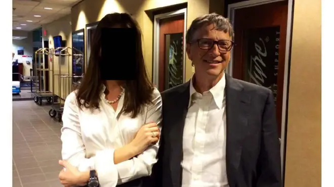 Bill Gates, con saco y camisa, posa junta una mujer con camisa blanca en un pasillo de un edificio