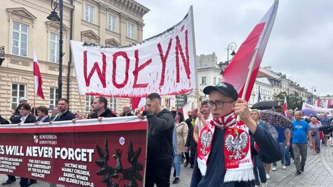 Вулична хода в центрі міста: учасники несуть польські прапори та великий банер із написом "Волинь". На передньому плані юнак із польським прапором і червоно-білим шарфом. Позаду видно натовп, банер англійською мовою про пам’ять жертв, а вдалині інший банер – із перекресленими українським гербом.
