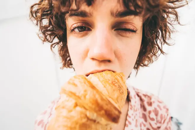 A woman bites down on a croissant, blinking one eye as she does so. The fish-eye shot exaggerates her face, the croissant and the bite. 
