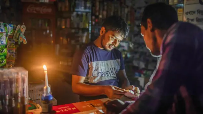 একটি দোকানে মোমবাতি জ্বালিয়ে কাজ করছেন একজন, সামনে আরেকজন দাঁড়িয়ে আছেন 