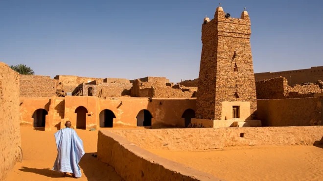 Un homme portant un grand boubou bleu ciel, marchant de dos, vers une mosquée.