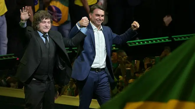 Javier Milei e Flávio Bolsonaro de mãos dadas para o alto.