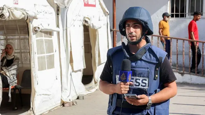 Anas al-Sharif stands next to a tent wearing a dark blue helmet and vest with "press" written on it, while he speaks into a microphone during a broadcast.