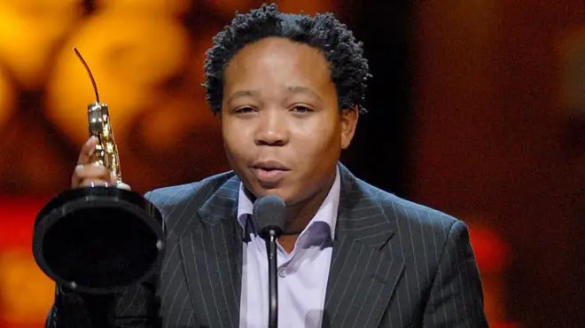 Presley Chweneyagae dey deliva a speech afta e win di Oscar Outstanding Performance by an Actor for a Leading Role for "Tsotsi". E dey lift im award up as e wear black stripe suit