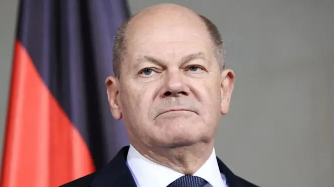 Foto do chanceler alemão Olaf Scholz, inexpressivobonus bet365 gratisfrente a uma bandeira vermelha e preta