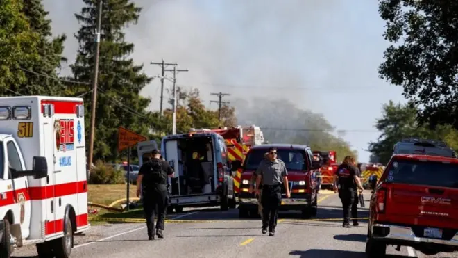 Agentes caminham em meio a carros de bombeiros e ambulância em via; ao fundo vê-se fumaça.