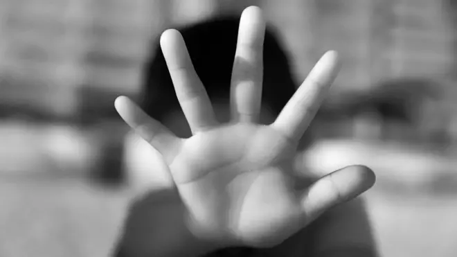 На чорно-білому знімку – піднята вперед дитяча рука у жесті "зупинись", що закриває більшу частину кадру. Фігура людини позаду розмита й нерозбірлива — видно лише темний силует.