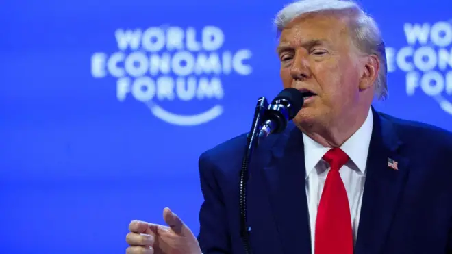 Доналд Трамп у тегет оделу са црвеном краватом док нешто показује десном руком