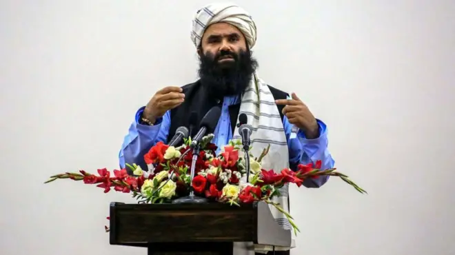 سراج الدين حقاني مرتدياً قميصاً أرزق وزياً أفغانياً وأمامه ورد أصفر وأحمر على منصة عليها مايكروفون