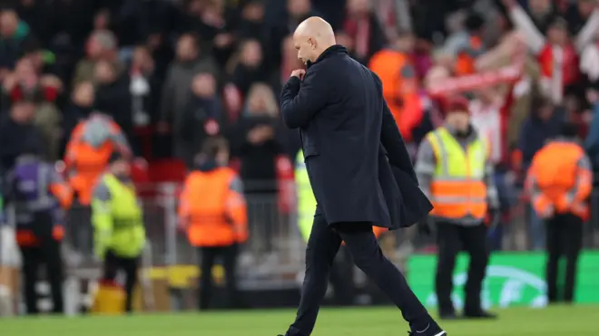 مدرب ليفربول، آرني سلوت يبدو واقفاً على أرضية الملعب، يرتدي "جاكيت" أسود اللون وبنطالاً أسود وحذاء رياضياً أسود.