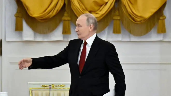 Торжественный зал с белыми стенами и большими желтыми драпированными шторами. В центре кадра — Путин в черном костюме с белой рубашкой и красным галстуком, который держит лист бумаги в левой руке и делает жест правой рукой.