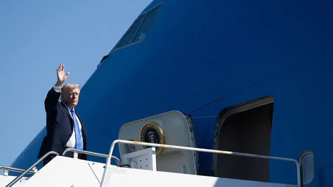 SArışın lacivert takım elbiseli bir adam büyük bir mavi uçağın kapısında el sallıyor. Uçağın kapısında Amerikan başkanlığının logosu var.