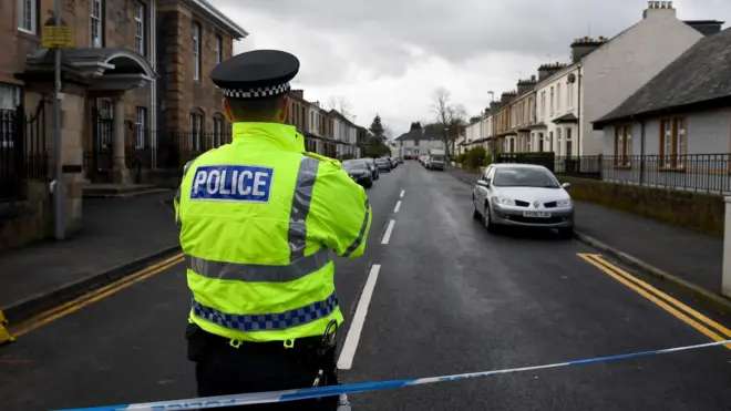 Um policial usando um colete de alta visibilidade atrás de uma fita de isolamento azul e branca que bloqueia uma rua residencial em Glasgow