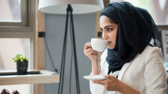 Une femme voilée boit du café dans une petite tasse blanche, son voile est noir, sa chemise est blanche et elle porte une veste de soirée blanche par-dessus.