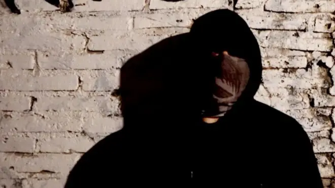 Мужчина в черной одежде с каплюшоном, лица почти не видно, т.к. оно закрыто платком. Он стоит у стены из светлого кирпича.