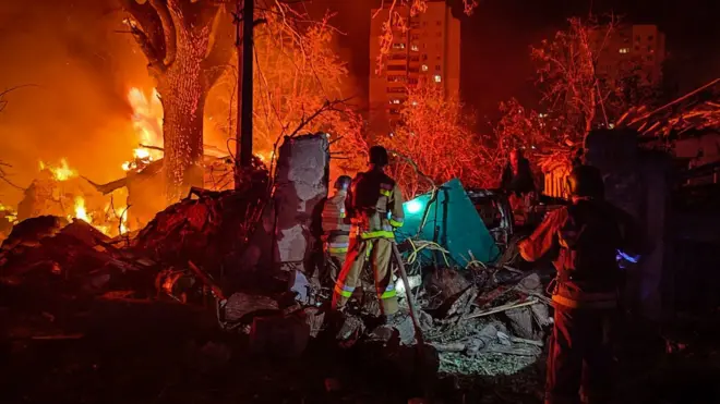 An emergency responder stands amid debris from a strike in Ukraine, lit up by a fire in the distance.