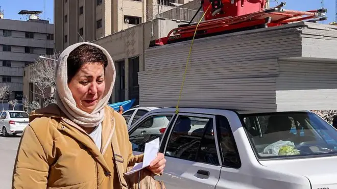 A woman appears to be crying as she walks on a street in Tehran. She has a light coloured headscarf and a tan coat on. She is holding a piece of paper in her hand and has a rucksack on her shoulder. A motorcyclist is behind her and cars line the street to her left.