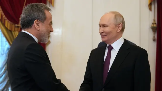 ダークスーツ姿の2人が笑顔で握手を交わしている。アラグチ氏はひげをたくわえている