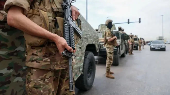 迷彩模様などの軍用車両が路上に並び、迷彩服姿の兵士たちが自動小銃などを手にしている。向こうから乗用車が走ってくる