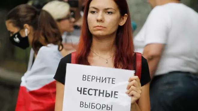 На фото, сделанном в августе 2020 года около посольства Беларуси в Киеве, крупным планом женщина в черной футболке, на плечах у нее темно-красные кожаные ремни от рюкзака. В руках у нее плакат "Верните честные выборы". На заднем плане еще одна участница акции, у нее на плечах бело-красно-белый флаг белорусской оппозиции 