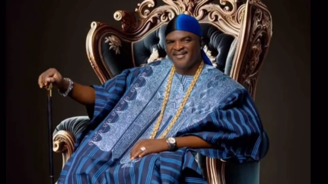 Obesere jòkòó lórí àga, wọ aṣọ olómi aro, mú ọ̀pá dání, wọ ṣéìnì sọ́rùn 