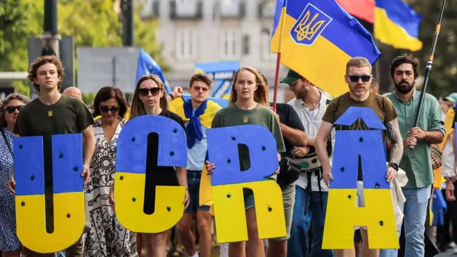 Мужчины и женщины на акции в поддержку Украины в Португалии держат в руках буквы большие U, C, R и A, окрашенные в синий и желтый цвета. На них футболки цвета хаки с украинскими трезубцами. На заднем фоне видны украинские флаги