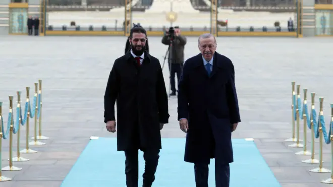 Erdoğan sağda, Şara solda, ikisi de siyah paltolu mavi bir halıda yürüyorlar, arkada bir fotoğrafçının flu görüntüsü var.