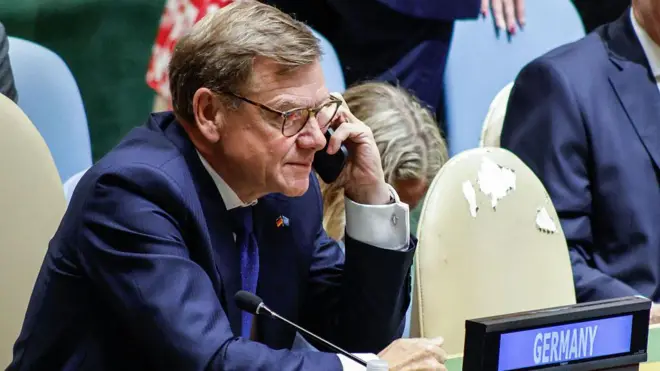 紺のスーツに青いネクタイを着けたヴァーデフール外相が、クリーム色の椅子に座り、左手で持ったスマートフォンを耳にあてている
