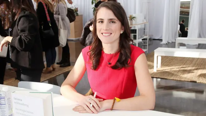 Tatiana Schlossberg with long brown hair and wearing a red short-sleeved dress, sits behind table where her book is stood up with hands crossed holding a pen