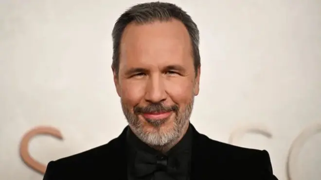 Вильнев хитро улыбается. Он одет в черный костюм с черным галстуком-бабочкой.